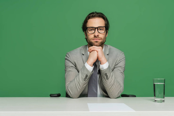 ραδιοτηλεοπτικός οργανισμός με γυαλιά ηλίου και γκρι κοστούμι κάθεται με σφιγμένα χέρια στο γραφείο απομονώνονται σε πράσινο - Φωτογραφία, εικόνα