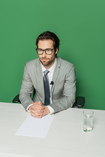 над головой просмотр новостей вещатель в очках и костюме сидя с сжатыми руками за столом изолированы на зеленый - Фото, изображение