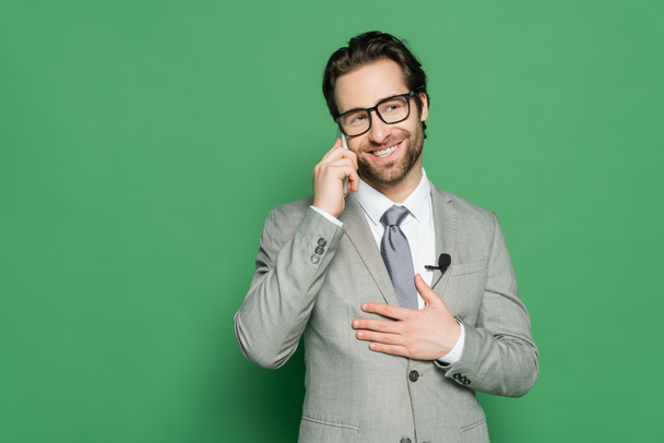 веселый репортер в очках и костюме говорить на смартфоне на зеленом фоне  - Фото, изображение