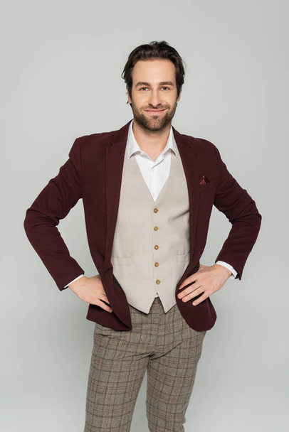 showman alegre en chaleco y chaqueta roja de pie con las manos en las caderas aisladas en gris  - Foto, imagen