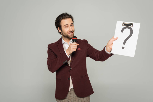 bearded host of event in formal wear talking in microphone while holding clipboard with question mark isolated on grey  - Photo, Image