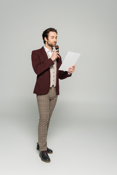 full length of bearded showman in official wear μιλώντας στο μικρόφωνο και κοιτάζοντας το χαρτί σε γκρι  - Φωτογραφία, εικόνα