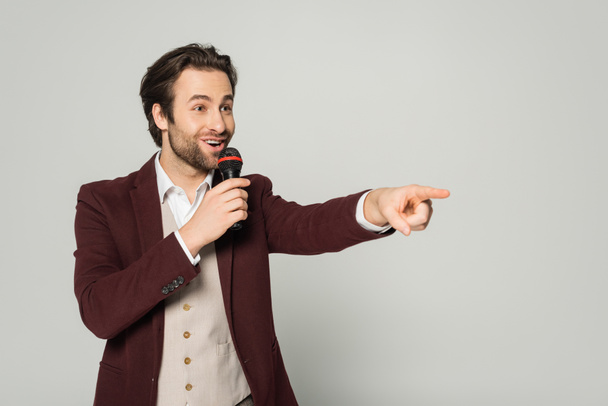 showman excitado en ropa formal hablando en micrófono y apuntando con el dedo aislado en gris  - Foto, Imagen