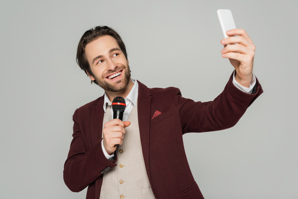 веселий шоумен бере селфі на смартфон, тримаючи мікрофон ізольовано на сірому
  - Фото, зображення