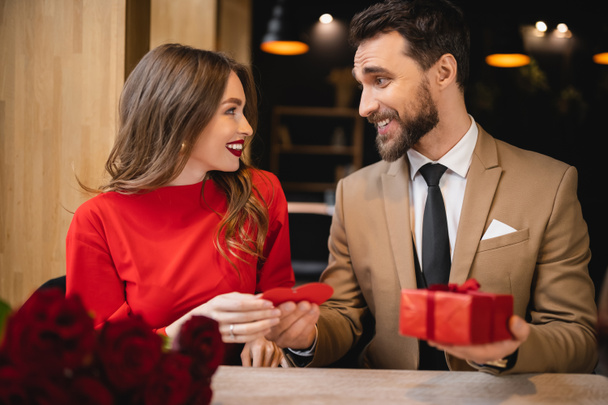 cheerful and bearded man giving red heart-shaped greeting card while holding present near happy girlfriend  - Foto, afbeelding