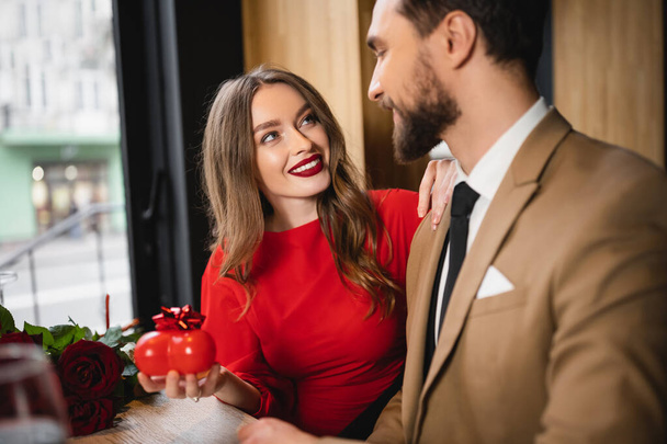 cheerful woman in red dress holding heart-shaped present with bow and looking at boyfriend on valentines day  - Photo, image