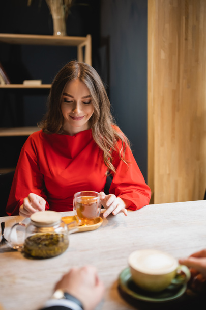 alegre joven mujer en vestido rojo mirando taza de vidrio con té verde cerca de novio   - Foto, Imagen