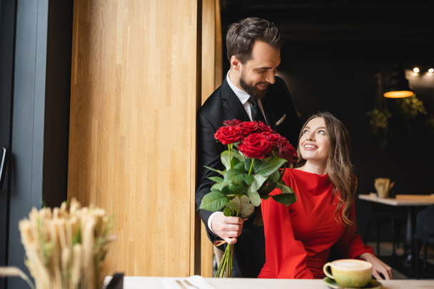 brodaty mężczyzna w garniturze trzymając bukiet czerwonych róż w pobliżu uśmiechniętej dziewczyny w Walentynki  - Zdjęcie, obraz