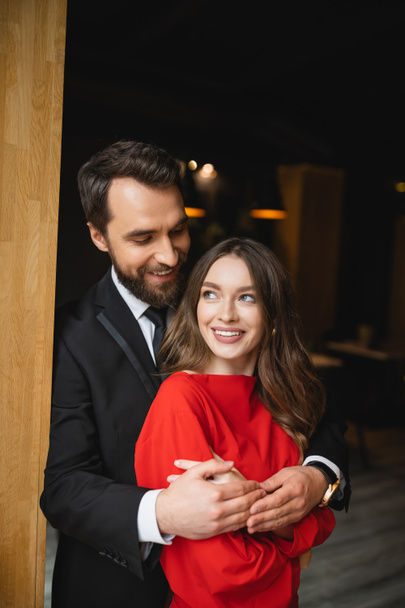 bärtiger Mann im Anzug umarmt glückliche Frau im roten Kleid am Valentinstag  - Foto, Bild