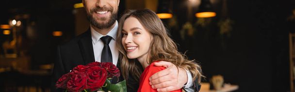 髭を生やした男は幸せなガールフレンドを抱きかかえてバレンタインデーに赤いバラの花束を  - 写真・画像