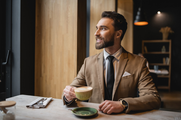 ευτυχισμένος γενειοφόρος άντρας με κοστούμι κρατώντας ένα φλιτζάνι καπουτσίνο στο καφέ  - Φωτογραφία, εικόνα