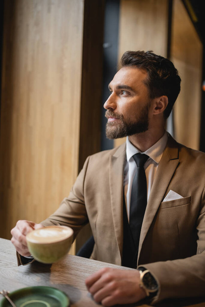 brodaty mężczyzna w garniturze trzyma filiżankę cappuccino i odwraca wzrok w kawiarni  - Zdjęcie, obraz