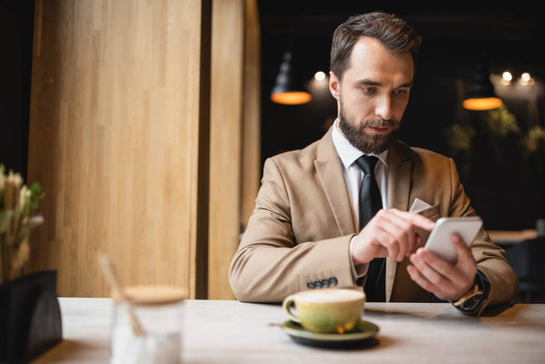 бородатый мужчина в костюме с помощью смартфона возле чашки капучино в кафе  - Фото, изображение