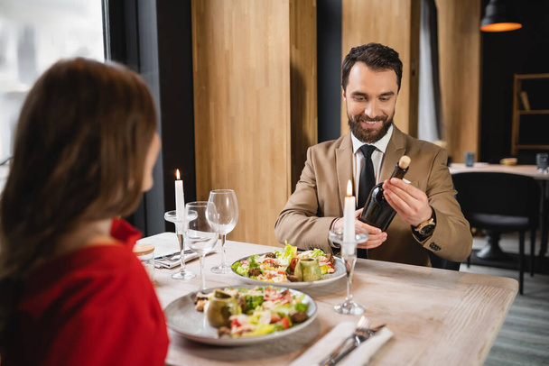uomo barbuto sorridente mentre tiene bottiglia con vino vicino fidanzata durante la cena festiva il giorno di San Valentino - Foto, immagini