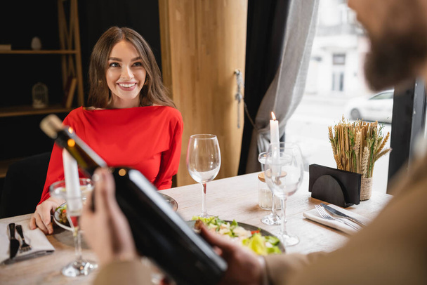 cheerful woman in red dress looking at man holding bottle with wine on valentines day - Photo, Image