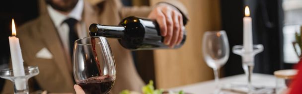 καλλιεργημένη άποψη του θολή άνθρωπος ρίχνει κόκκινο κρασί σε ποτήρι της φίλη κατά τη διάρκεια του δείπνου την ημέρα του Αγίου Βαλεντίνου, πανό  - Φωτογραφία, εικόνα