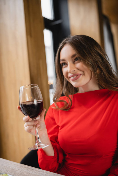 Kırmızı şarapla bardak tutan ve restoranda gülümseyen mutlu genç bir kadın.  - Fotoğraf, Görsel