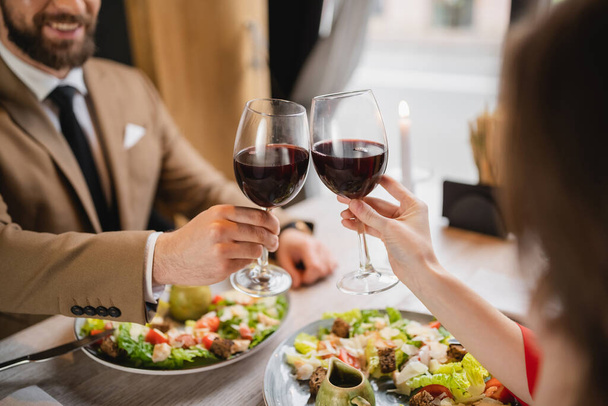 Sevgililer Günü 'nde kutlama sırasında yemek yakınında kırmızı şarapla kadeh tokuşturan çift manzarası.  - Fotoğraf, Görsel