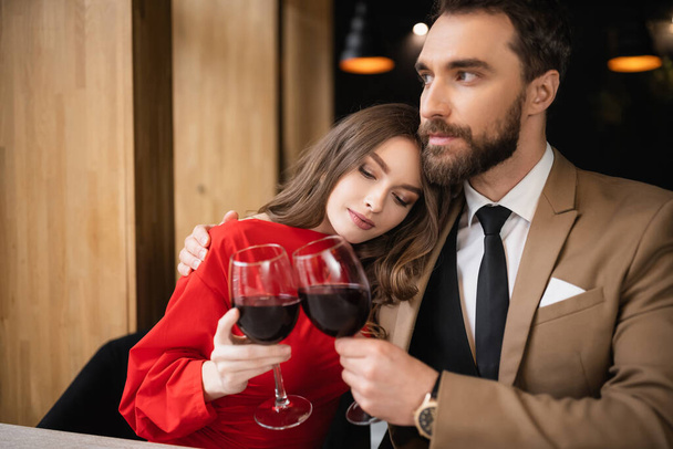Genç kadın ve sakallı adam sevgililer gününde kutlama sırasında kırmızı şarapla kadeh tokuşturuyorlar.  - Fotoğraf, Görsel