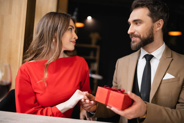 vousatý muž ve formálním oblečení držení dárek poblíž usmívající se žena v červených šatech na Valentýna  - Fotografie, Obrázek