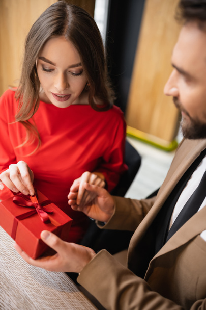 szakállas férfi formális viselet gazdaság ajándék közelében fiatal nő piros ruhában Valentin-napon  - Fotó, kép