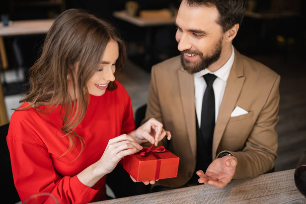 бородатый мужчина в официальной одежде с подарком в то время как счастливая женщина в красном платье тянет ленточку на День Святого Валентина  - Фото, изображение