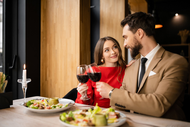 joyeux jeune femme et barbu homme cliquetis verres avec du vin rouge lors de la célébration sur la Saint-Valentin  - Photo, image