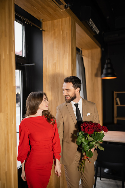 uomo barbuto che tiene rose rosse mentre guarda la fidanzata allegra in abito il giorno di San Valentino - Foto, immagini