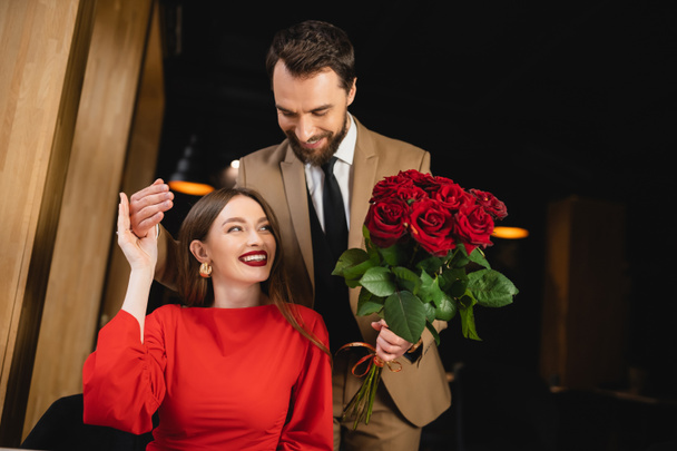 bearded man in formal wear holding bouquet of red roses near cheerful woman on valentines day  - Foto, afbeelding