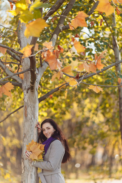 una hermosa chica con el pelo oscuro, un abrigo húmedo y una bufanda púrpura sostiene una carta. fondo borroso y hojas borrosas visibles arriba - Foto, imagen