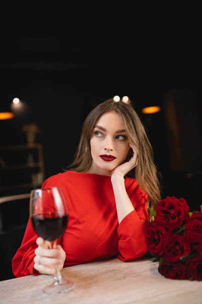 dromerige jonge vrouw met glas rode wijn in de buurt van rozen op Valentijnsdag - Foto, afbeelding