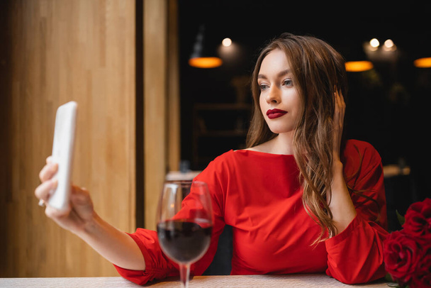 pretty young woman with red lips taking selfie near glass of wine and roses on valentines day - Photo, Image