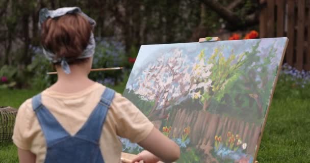 Chica creativa está pintando un hermoso cuadro en el jardín, sosteniendo una paleta y un pincel, utilizando herramientas para crear un lienzo. Concepto de artistas y obras. - Imágenes, Vídeo