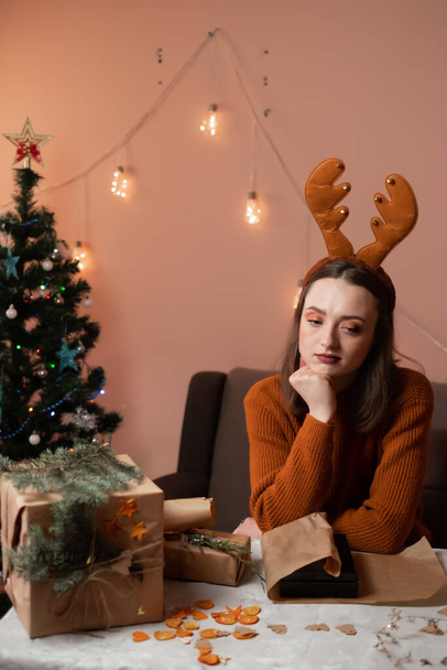 щаслива молода жінка готується до різдвяних і пакувальних подарунків, у дівчини новорічні роги на голові, на задньому плані ялинка і вогні, концепція підготовки до нового року, веселий різдвяний, новий рік 2023
 - Фото, зображення