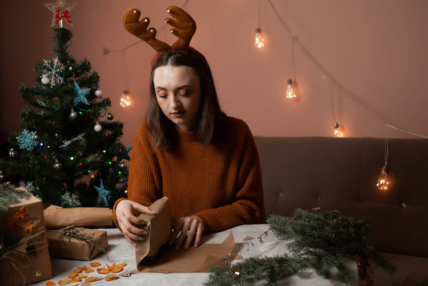 a happy young woman is preparing for christmas and packing gifts, the girl has New Year's deer antlers on her head, a Christmas tree and lights are in the background, the concept of preparing for the new year, merry christmas, new year 2023 - Photo, Image