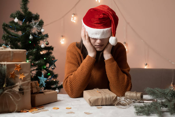 Yılbaşı şapkası takmış yorgun genç bir kadın gözlerini çekti hediye paketi siparişi aldı, arka planda bir Noel ağacı ve ışıklar, yeni yıla hazırlık konsepti, mutlu noeller, 2023 - Fotoğraf, Görsel