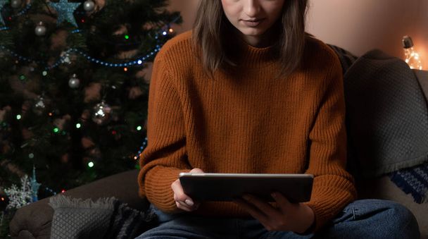 Eine junge Frau hält ein Telefon, ein Tablet in der Hand und erfüllt telefonisch eine Bestellung, nimmt eine Bestellung für Geschenkverpackung entgegen, im Hintergrund einen Weihnachtsbaum und Lichter, das Konzept der Vorbereitung auf das neue Jahr, frohe Weihnachten, Neujahr 2023 - Foto, Bild