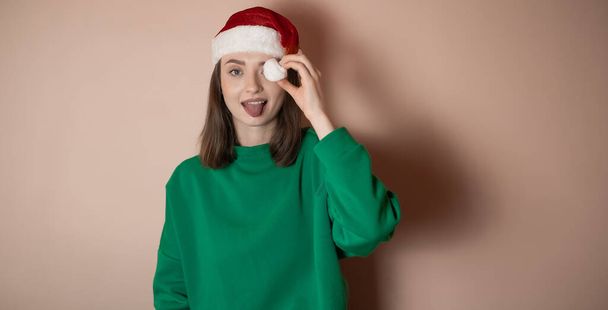 mujer joven en un sombrero de Año Nuevo, una mujer muestra su lengua, cubrió su ojo con un pompón de un sombrero, el concepto de la preparación para el nuevo año, Feliz Navidad, año nuevo 2023, chica feliz, fondo monocromo, llave de cromo - Foto, Imagen