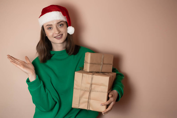 Весело удивлена, что молодая женщина носит рождественский свитер Санта шляпа позирует указывая руки в сторону указывают на рабочее пространство области изолированы на простой пастельной светло-зеленый фон. С Новым 2023 годом! - Фото, изображение