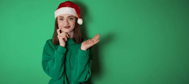 Merry surprised young woman wear xmas sweater Santa hat posing pointing hands arms aside indicate on workspace area isolated on plain pastel light green background. Happy New Year 2023 holiday concept - Photo, Image