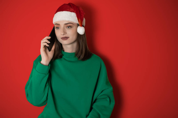 Девушка в новогодней шляпе разговаривает по телефону на обычном фоне, держа в руке телефон, заказывая ужин, концепцию новогодних выходных 2023 года, счастливого Рождества, 2023 года, праздников - Фото, изображение