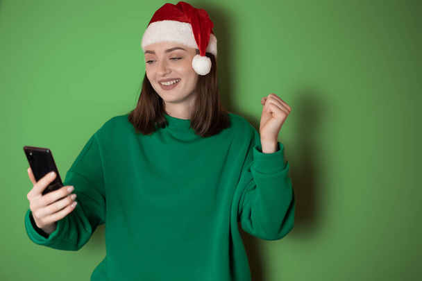 Cheerful young woman dressed in a green sweater and santa hat, holding a mobile phone in her hand, making a winner gesture, isolated on a green background. Happy new year 2023 holiday concept - Photo, Image