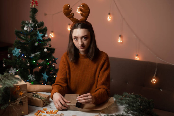 Mutlu bir genç kadın Noel için hazırlanıyor ve hediyeler paketliyor, kızın kafasında yeni yıl geyik boynuzları var, arka planda bir Noel ağacı ve ışıklar, yeni yıla hazırlık konsepti, mutlu noeller, yeni yıl 2023 - Fotoğraf, Görsel