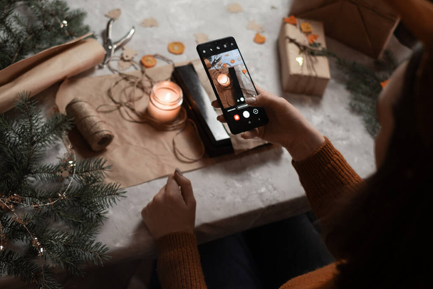 молодая женщина держит в руке телефон, планшет и выполняет заказ по телефону, принимая заказ на подарочную упаковку, на заднем плане новогодняя елка и огни, концепция подготовки к новому году, с Рождеством Христовым, Новый 2023 год - Фото, изображение
