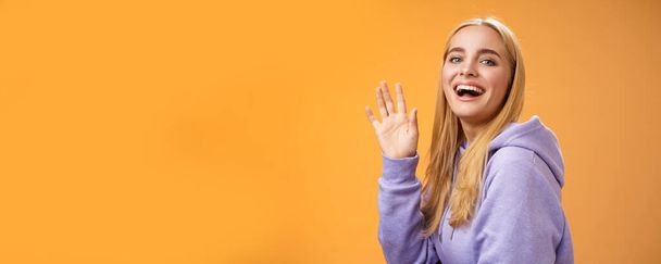 Waist-up friendly lucky cute blond girl waving palm happily smiling turning camera greet friend say hello hi see familiar person welcoming newcomers joyfully, standing orange background. - Photo, Image
