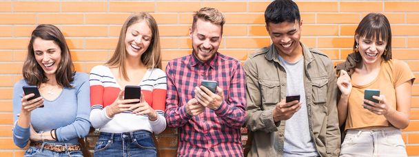 多文化の友達と一緒に大学のキャンパスの壁にスマートフォンを見て楽しんでいる横のバナー-携帯電話で中毒の若者 - 写真・画像