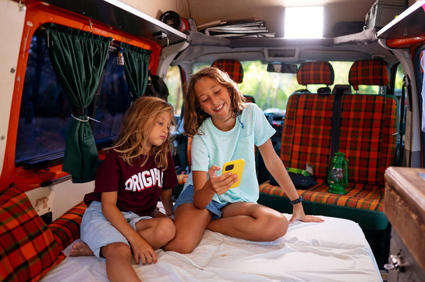 Minibüste kamp yaparken akıllı telefonu kullanan iki kardeş. Van yaşam konsepti. - Fotoğraf, Görsel