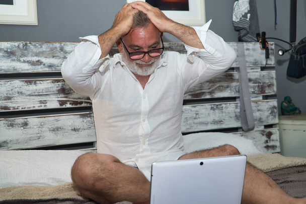 Homme mûr, cheveux gris et barbu, portant des lunettes, en pyjama, assis sur le lit, désespéré avec les mains sur la tête avec son ordinateur portable. Concept solitude, retraite, pensionné, technologie. - Photo, image