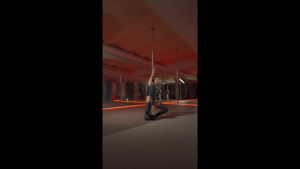 Joven mujer morena en forma delgada con el pelo largo en tacones altos está bailando en un poste en el club con luces rojas del piso. Imágenes verticales 4K. - Metraje, vídeo