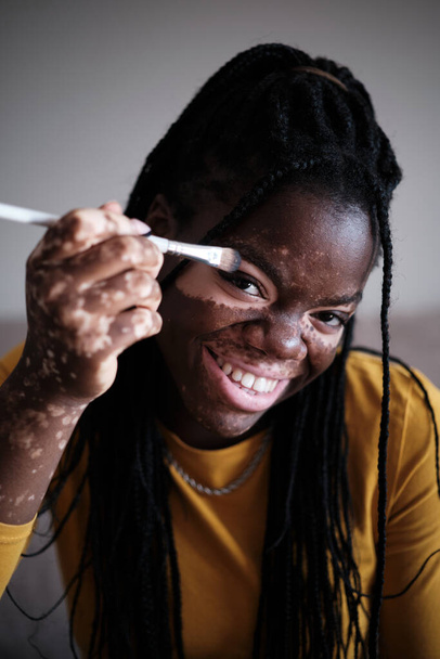 Femme afro-américaine joyeuse aux cheveux foncés souriant et regardant la caméra tout en faisant du maquillage avec une brosse près des yeux - Photo, image